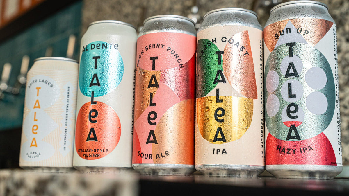 Five cans of Talea beer with colorful labels on a wooden surface.